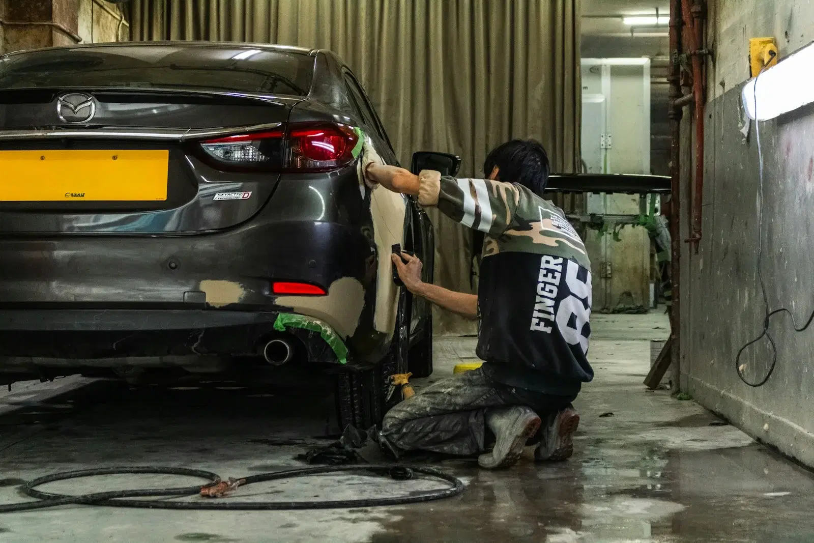 Technician hand-preparing a rear quarter panel with exposed body filler and masking tape