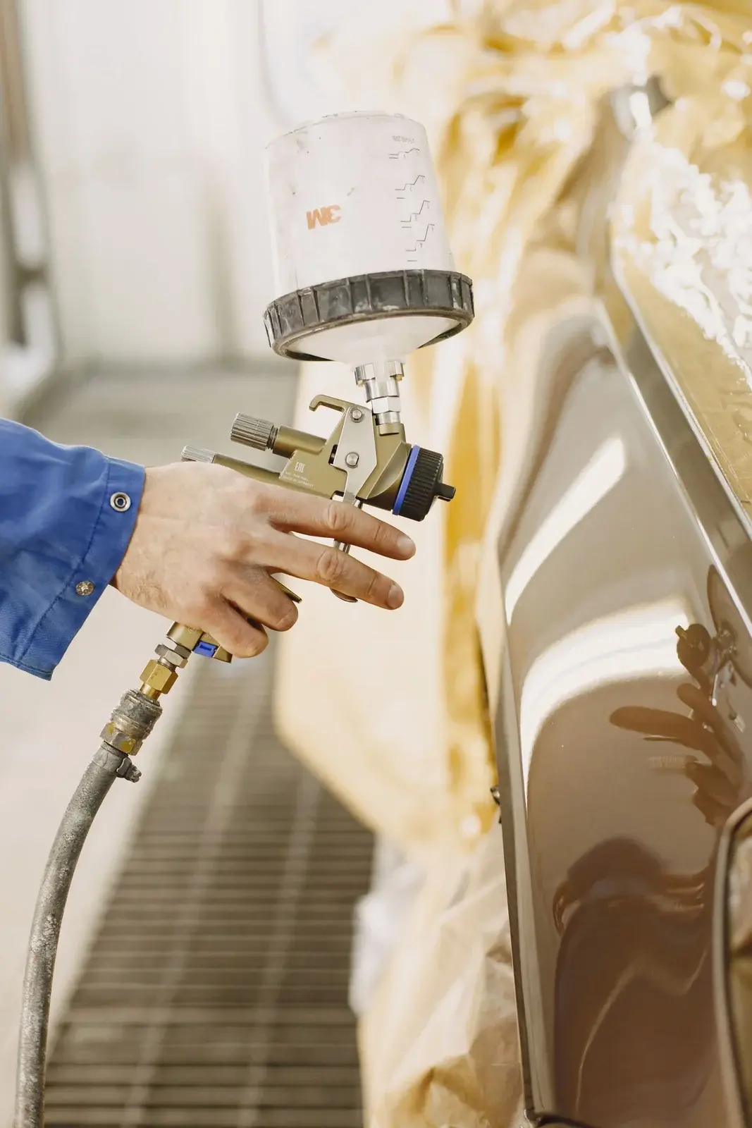 Paint spray gun directed at a silver body panel with adjacent areas masked for refinishing